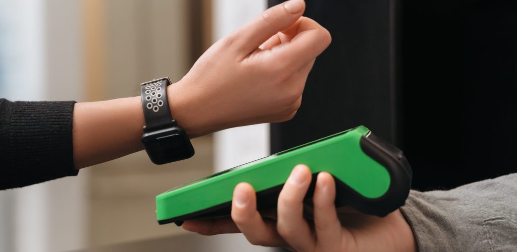 Closeup of modern lady paying via smartwatch while making purchase in shop. Woman customer using hand watch for contactless payment via payment terminal on counter in store. NFC technology concept
